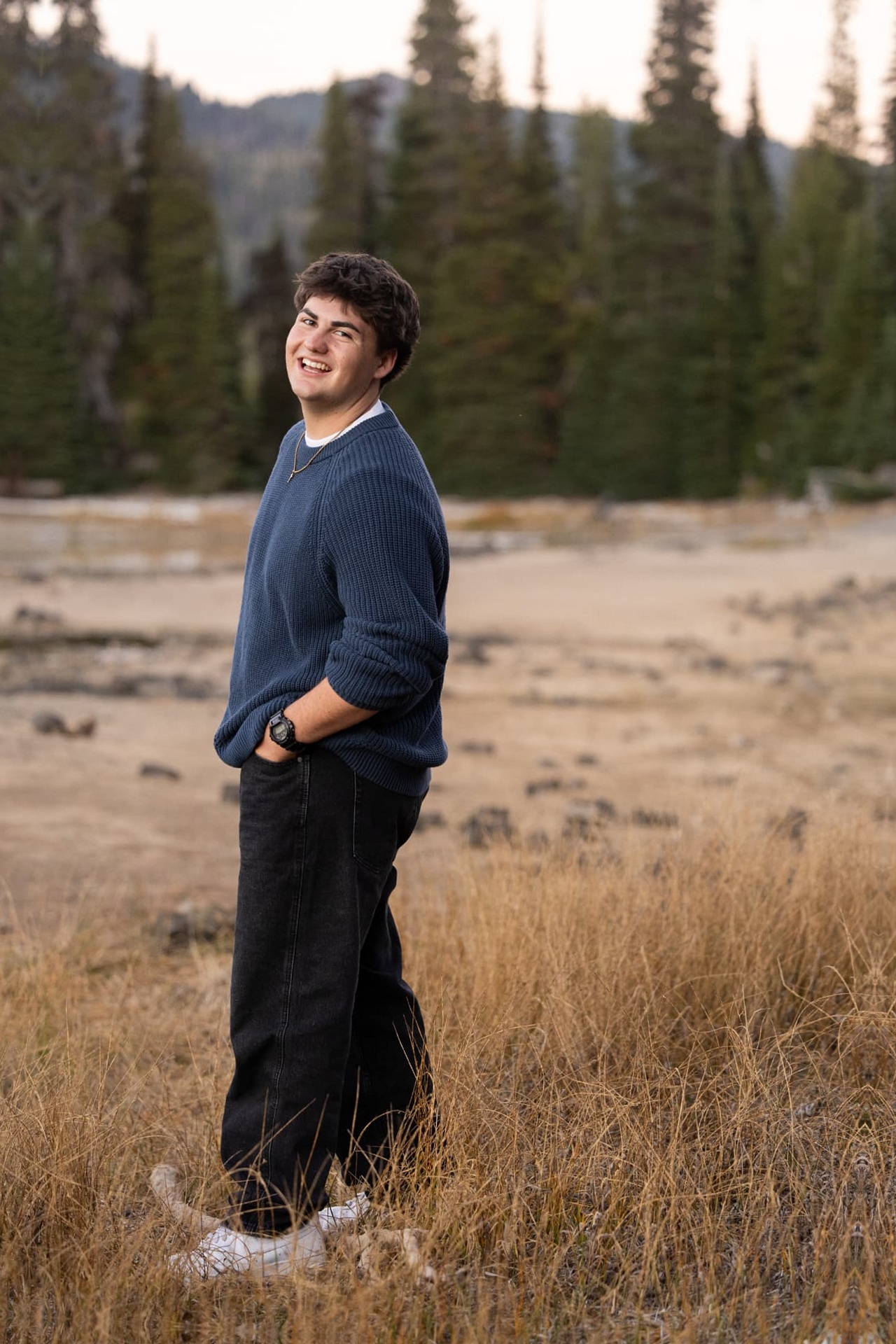 Senior guy in a navy sweater laughing in dry grass below the evergreens