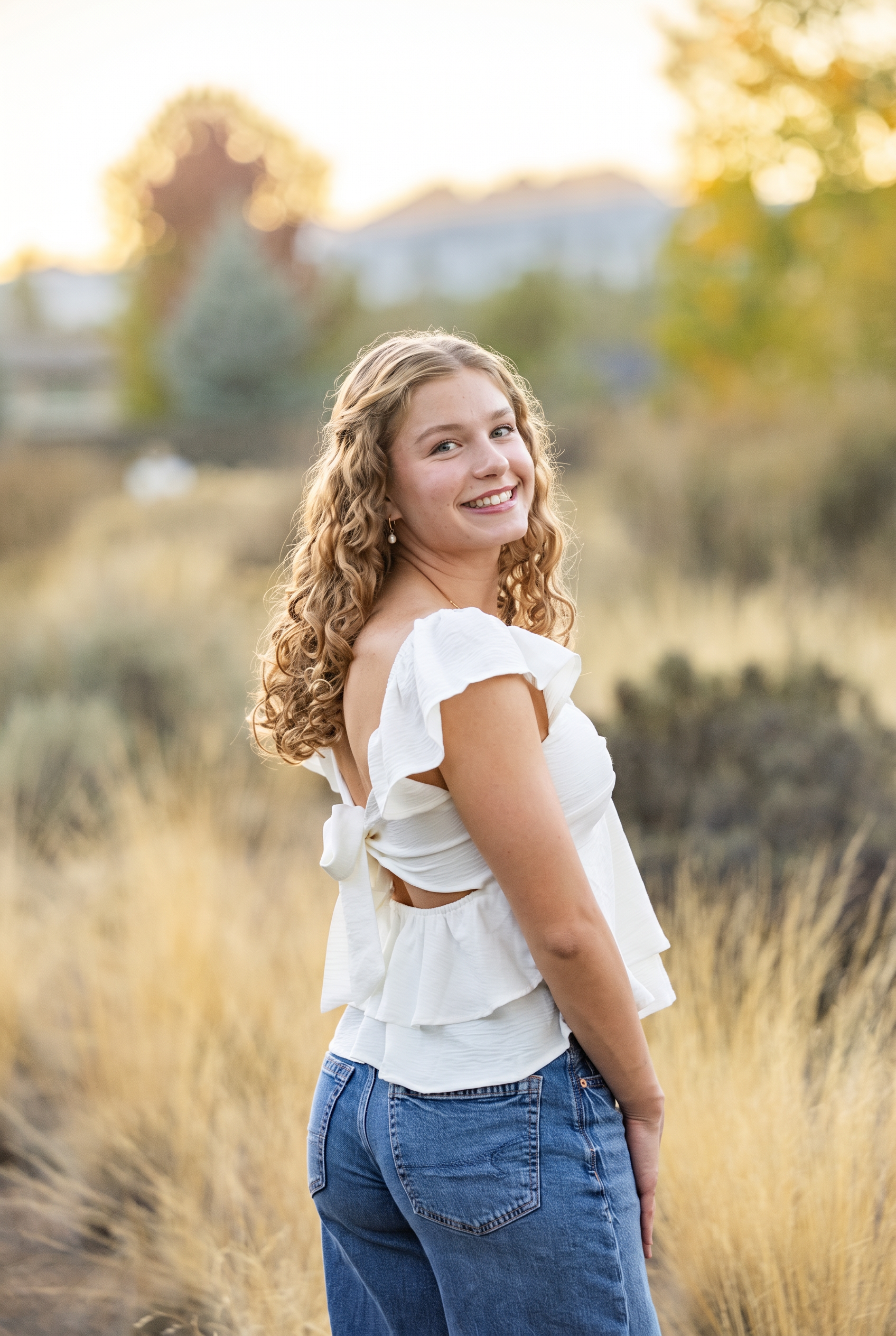 Senior girl in a white top and jeans looking over her shoulder in a golden meadow